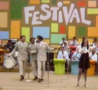 Film still of a stage with performers and the word "FESTIVAL" on a colorful backdrop