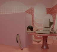 Woman's head and limbs emerge from a pink box to type on a computer in a surreal video still