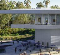 Rendering of elevated gray building with floor to ceiling windows, art, and visitors, and picnic tables in the foreground 